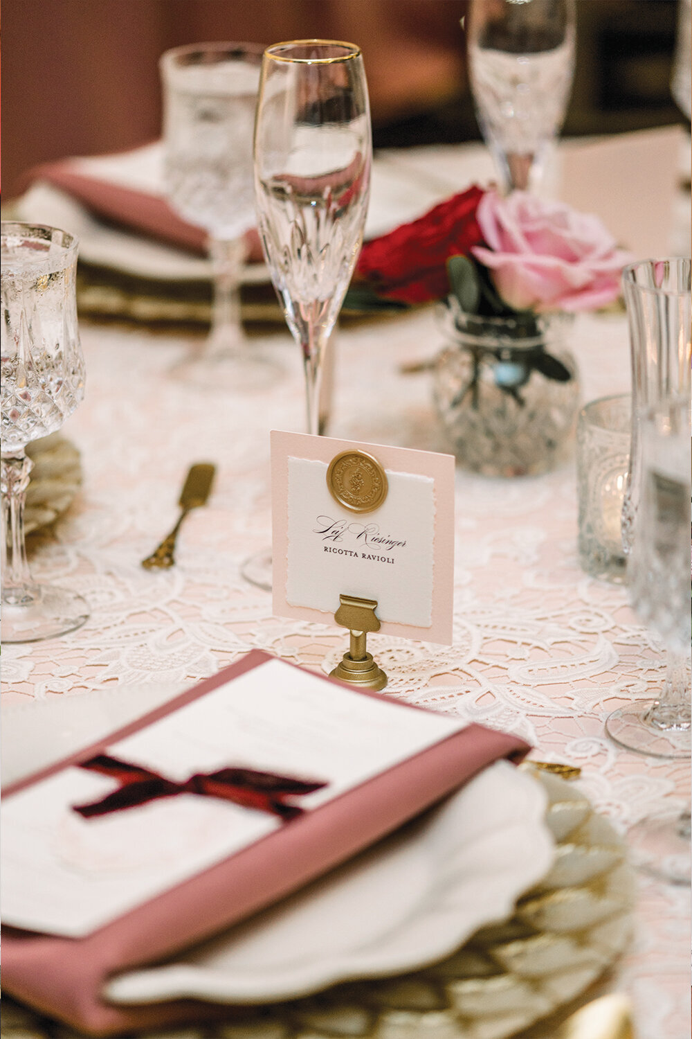 Chic place setting featuring gold chargers, dusty rose napkins wrapped around the charger, crystal glasses, and wax seal adorned place cards for a 50th wedding anniversary celebration at The Northern Hotel in Billings, MT.