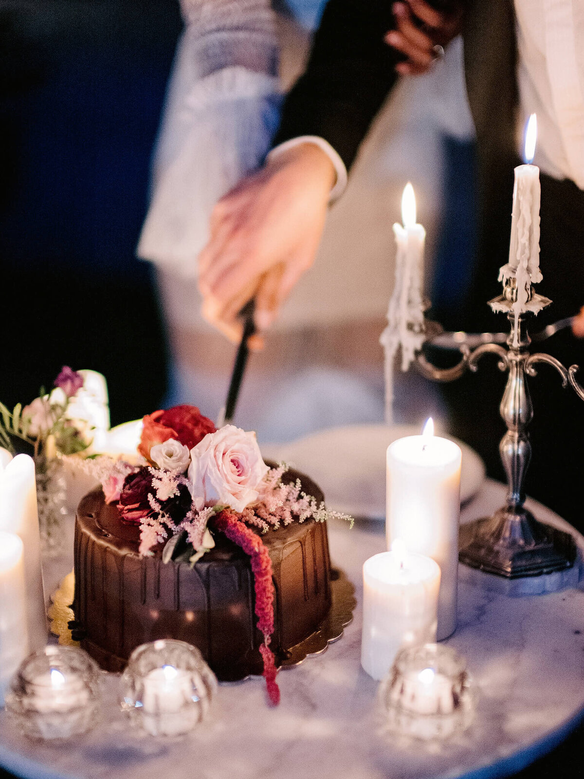 A chocolate cake with pink and red flowers on top, is on a table with candles around it, with two hands cutting it.
