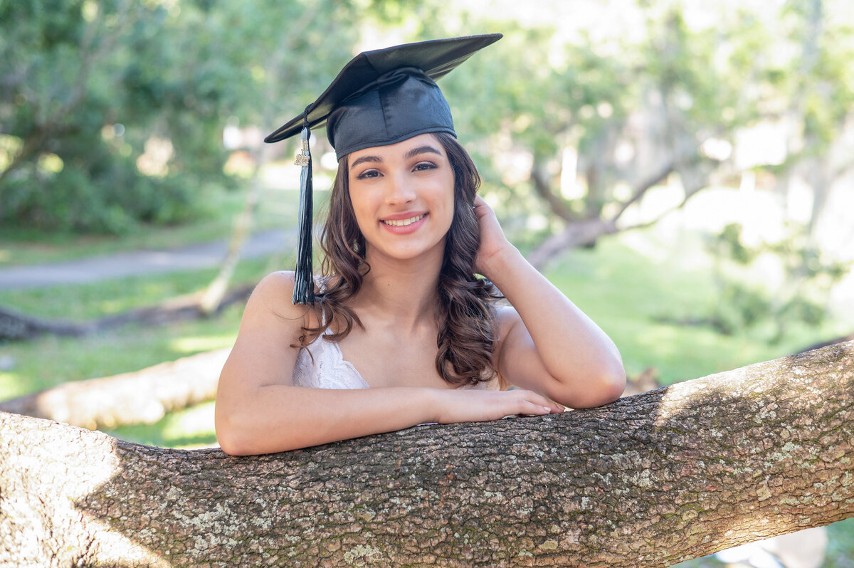High school senior girl wearing cap leans against a tree branch.