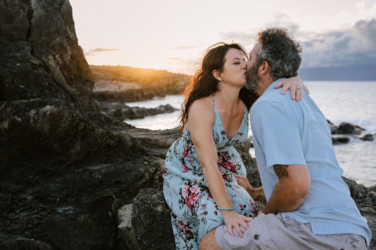 A man and a woman kiss on a rocky shore of Maui with the sunset in the background.