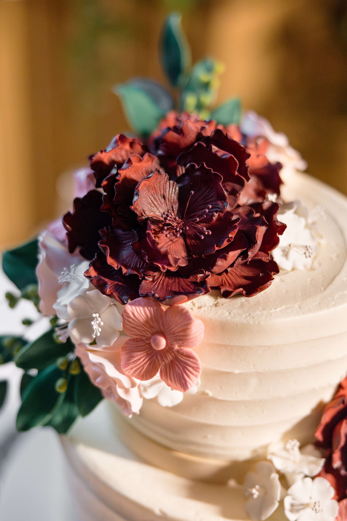 A close-up of a tiered cake decorated with intricate burgundy and pink flower designs made of icing or fondant, set against a warm, blurred background. The flowers include large burgundy blooms and smaller pink and white blossoms with green leaves.