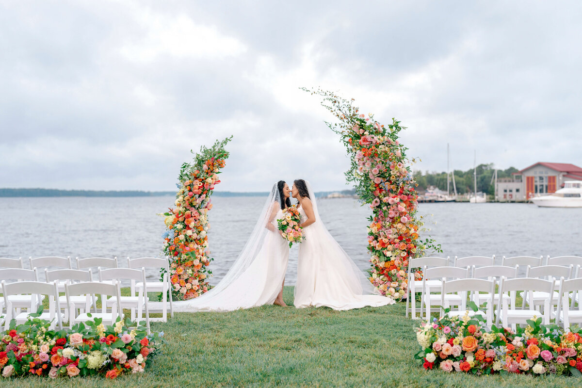 Two brides in white wedding dresses stand on a grassy area by the water, sharing a kiss. They are surrounded by colorful floral arches. White chairs are arranged in rows, with boats and a building visible in the background.