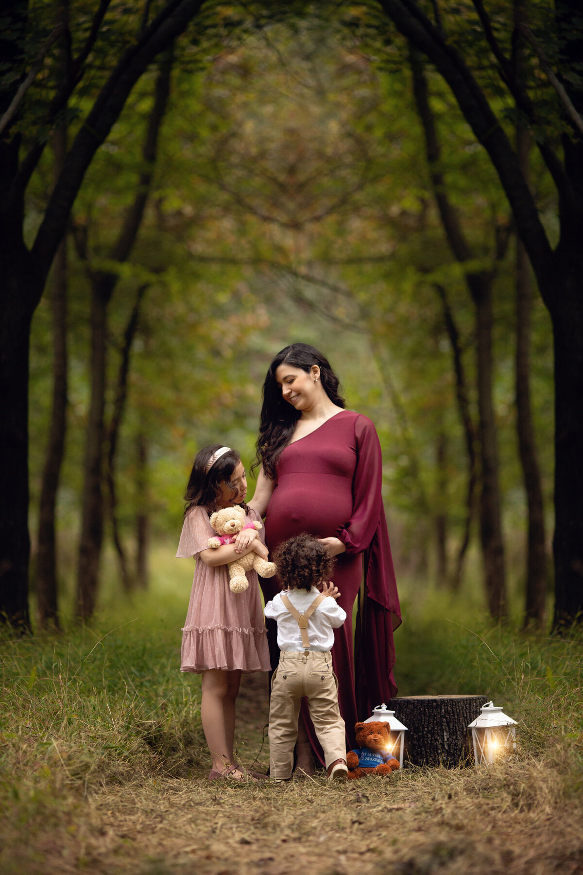 A pregnant mother in a maroon maternity dress plays with her toddler son and daughter in a forest trail for a New Jersey Maternity Photographer