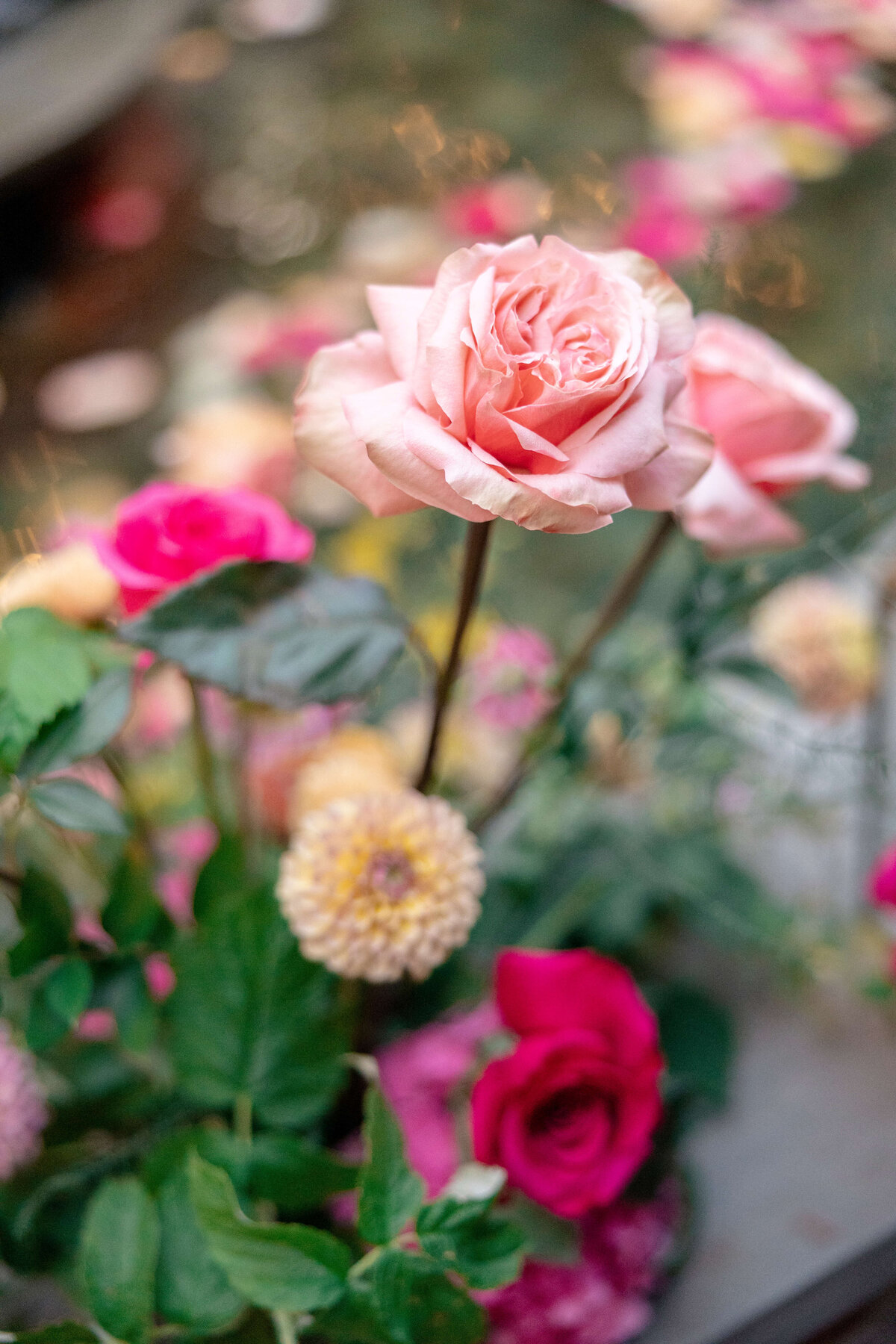 A close-up of a floral arrangement featuring a prominent pink rose surrounded by other flowers in soft pink and cream hues. The background contains more scattered blossoms, creating a subtle, blurred effect.