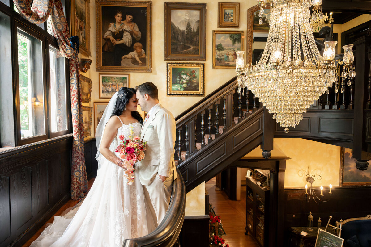 A bride in a white gown and groom in a light-colored suit stand on a grand staircase adorned with art and a chandelier. They gaze into each others eyes, with the bride holding a bouquet of pink and red flowers.