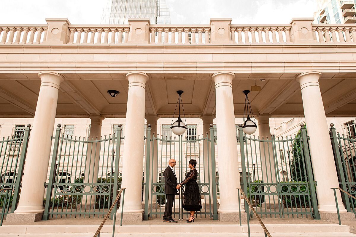 The bride and groom stand amongst a colonnade with long ivory fluted columns and decorative iron gates. They face each other holding hands. The groom is wearing a black suit and the bride is wearing a tea length black dress with long sheer sleeves.