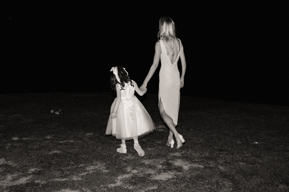 A woman in a backless dress and a young girl in a white dress walk hand in hand at night during an event in Davenport, viewed from behind.