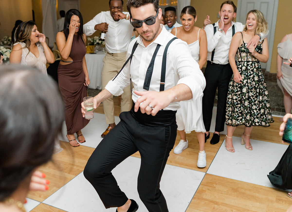 Groom grabs his sunglasses and hits the dance floor. Black tie groom with bowtie undone on shirt. Wedding flash photography.