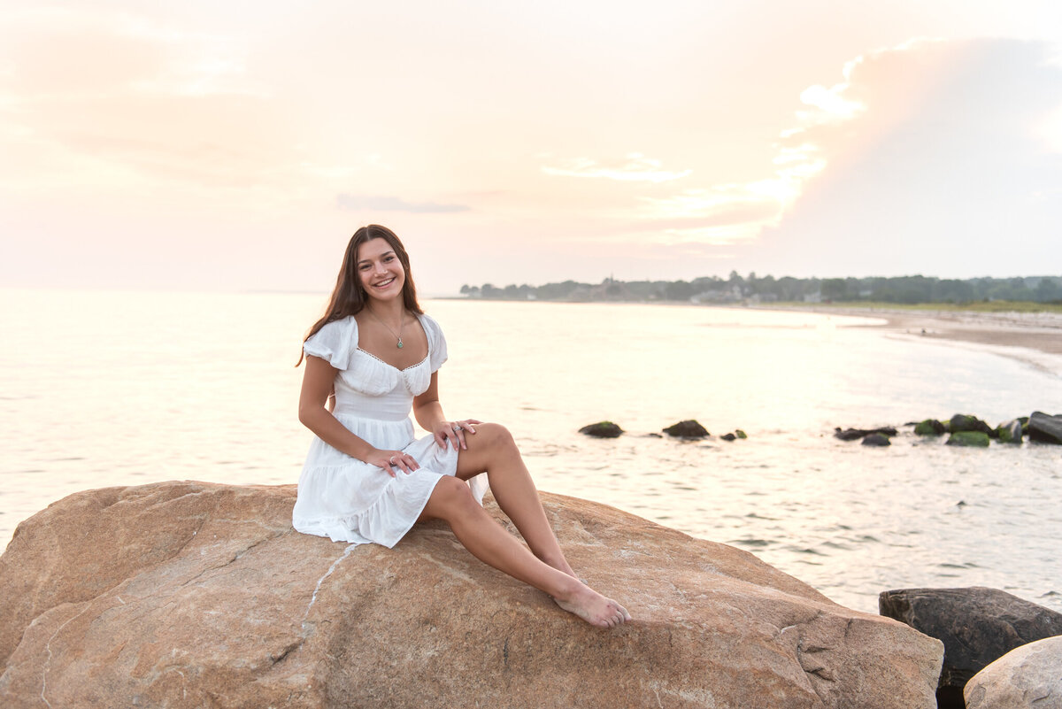 High school girl in white dress sitting on rock at beach during sunset