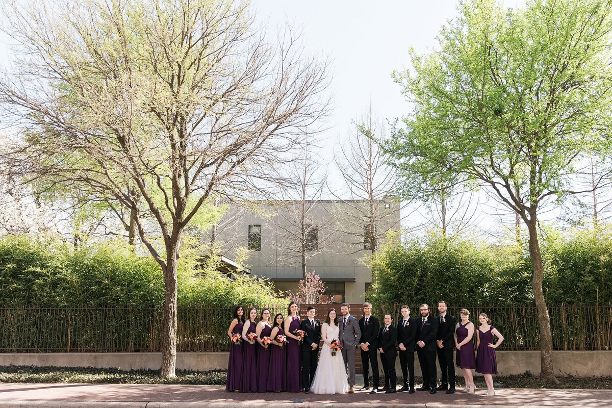 An outdoor portrait of a bride, groom, and their wedding party after their wedding ceremony in Plano, Texas. The bride is in the center on the left and is wearing an intricate, short sleeve, white dress and is holding a bouquet. The groom is in the center on the right and is wearing a grey suit with a boutonniere. The wedding party flanks them on either side and has members wearing either a black suit with a purple tie and boutonniere or a purple dress with a bouquet.