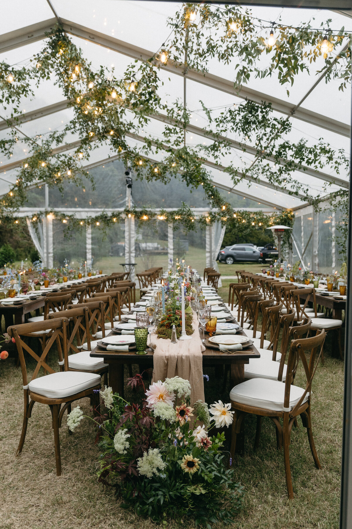 Dreamy wedding reception tables set beneath a clear-top tent with hanging greenery installations, twinkle lights, long wooden tables, and lush garden-style centerpieces designed by a Northwest Arkansas wedding florist for a romantic outdoor celebration.