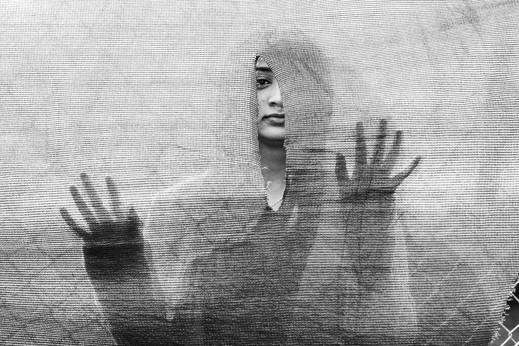 Girl stands behind a mesh barrier for a  portrait in Alameda, CA