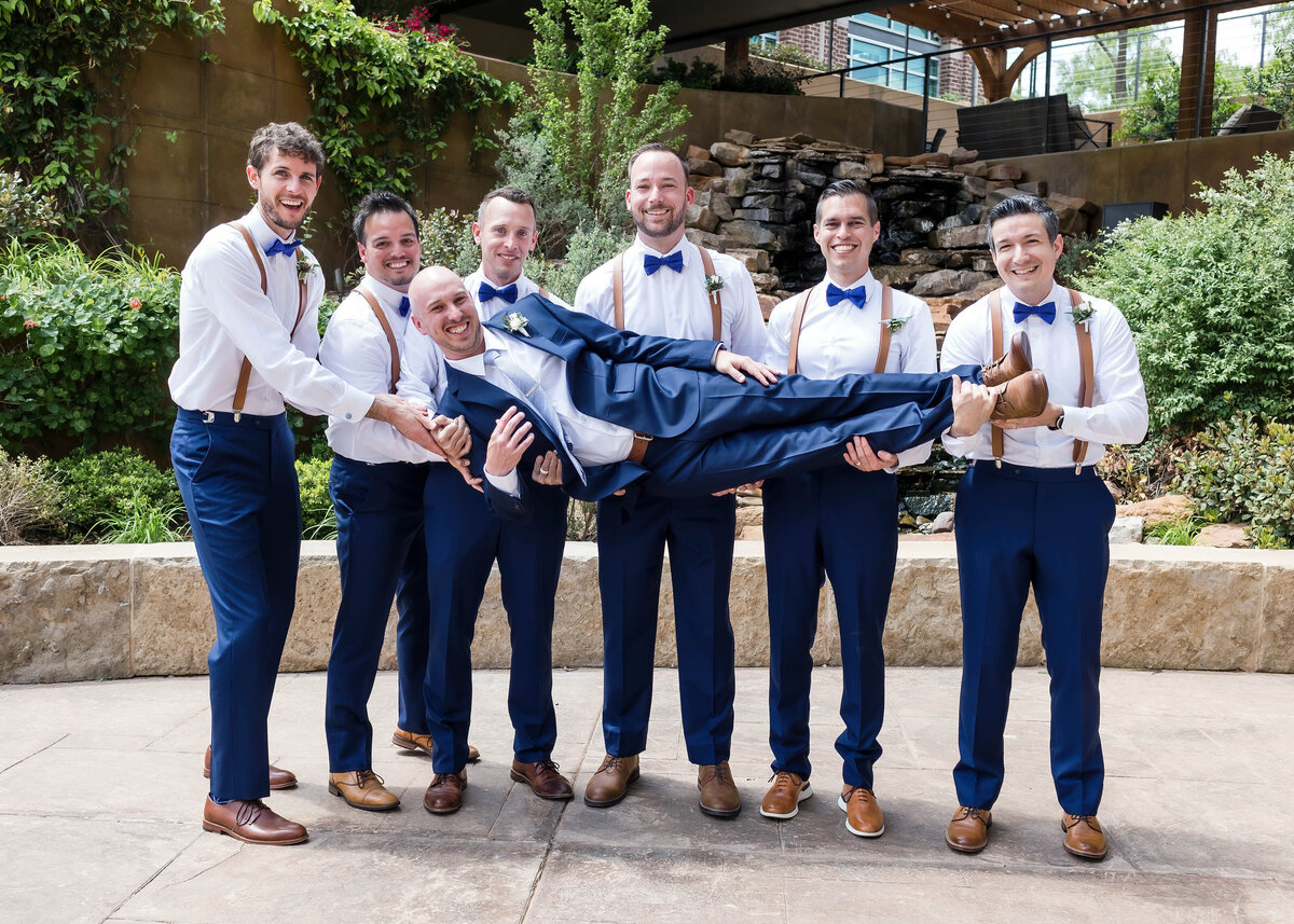 Groom and groomsmen posed in the Willows Event Center courtyard.  Groomsmen are wearing blue suit pants, white shirt, blue bowties and brown suspenders and groom is wearing a blue suit.  Groomsmen are holding the groom and all are looking at the camera.
