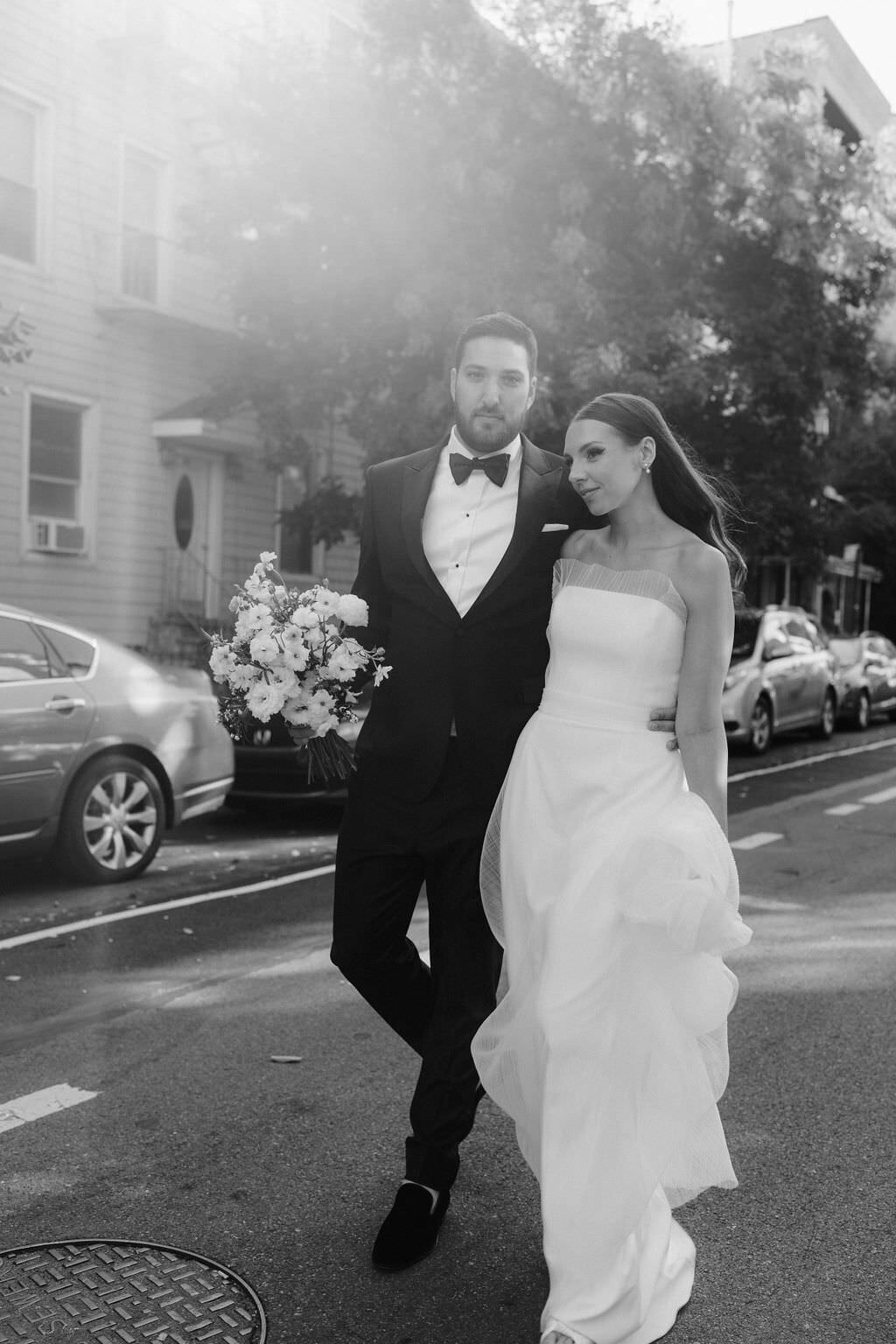 black and white photo of a groom holding bouquet with one hand while the other is wrapped around a bride as they walk down a street