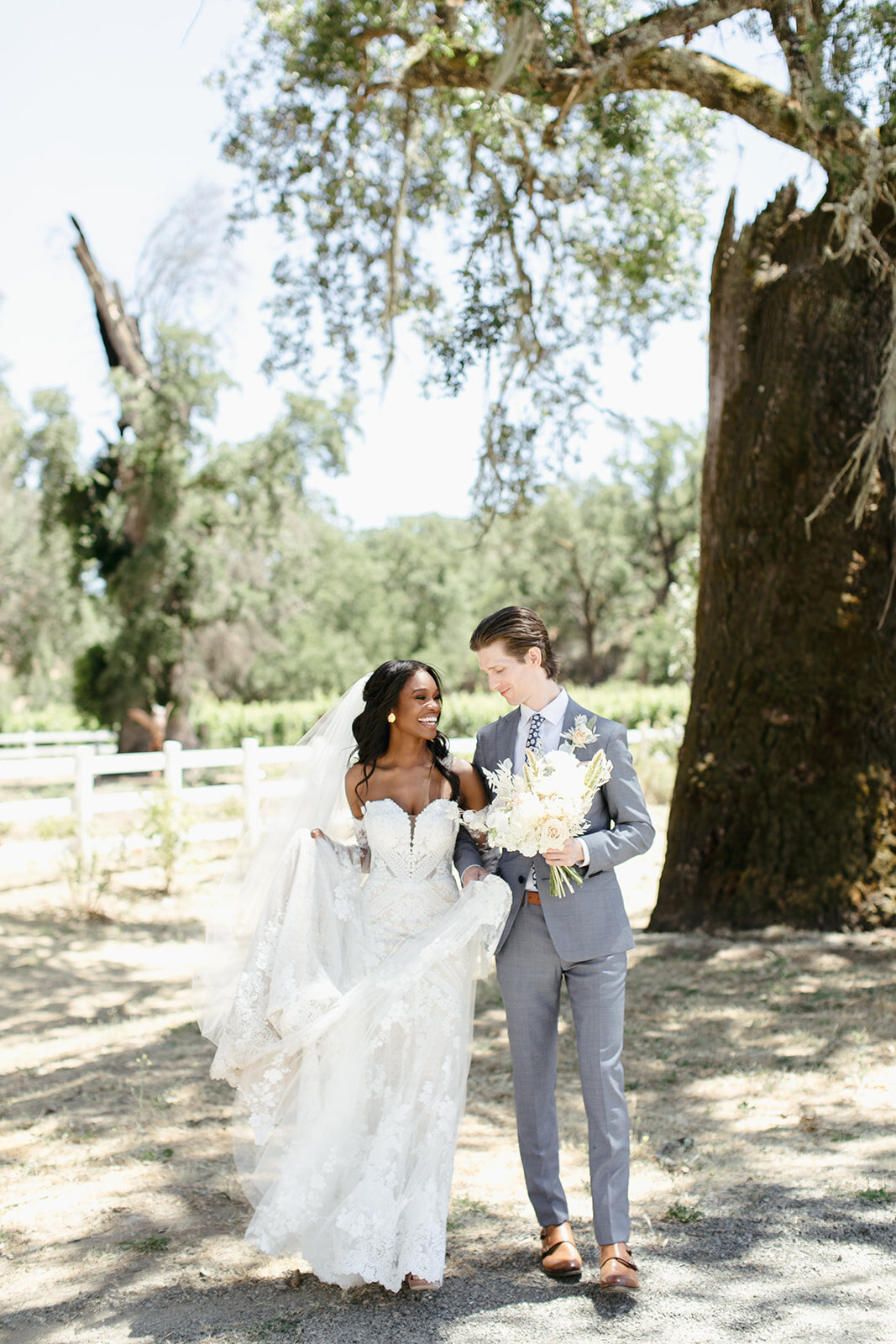 Bride and groom laugh as they walk together under large tree in Calistoga, California.