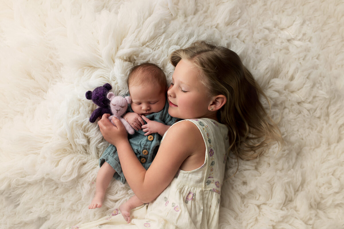 Portrait of sleeping infant with older sister snuggling on a fuzzy white rug in our Metro-Milwaukee photo studio.