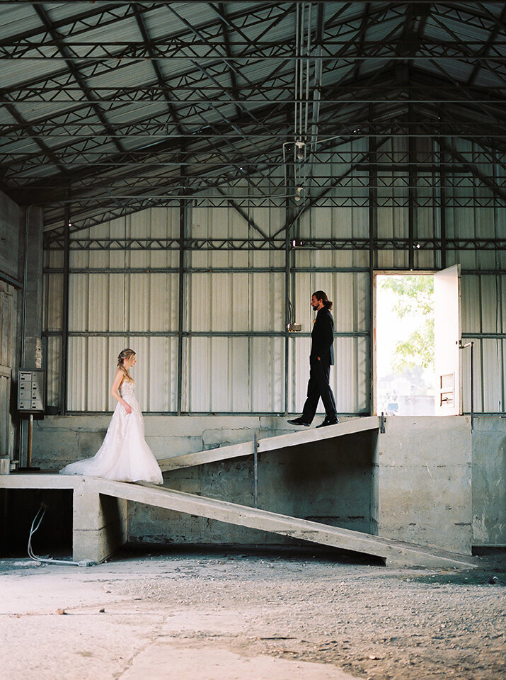 Bride and groom wearing a black tuxedo and white wedding gown walking down concrete slabs in an industrial warehouse.