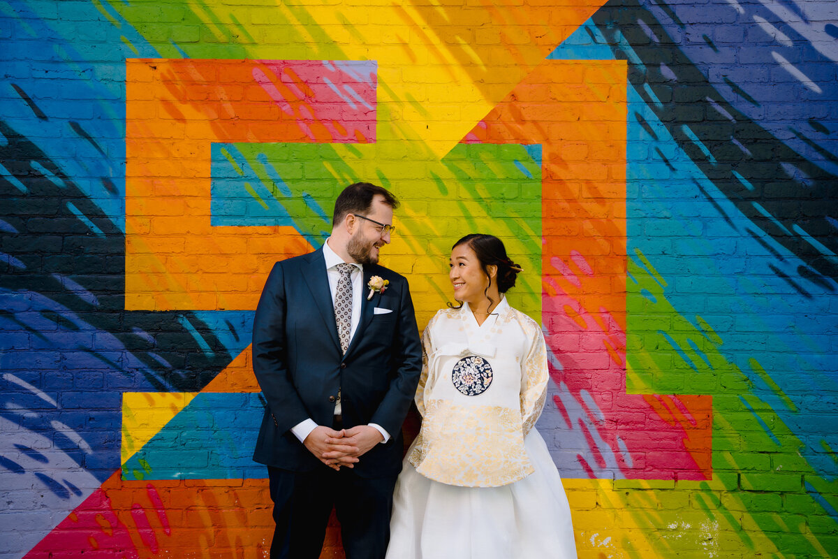 A bride and groom standing next to each other looking at each other while standing in front of a brightly painted wall.