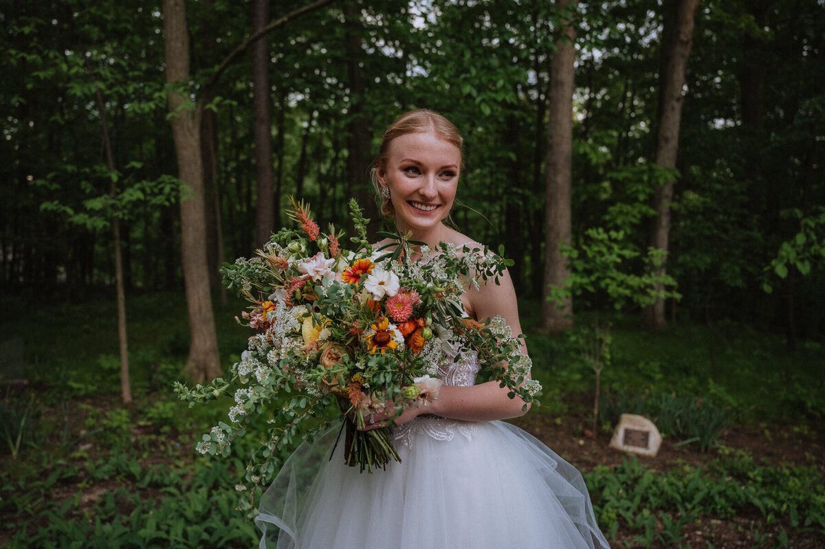 Bride in white ballgown style wedding dress holding colorful bouquet of flowers in the woods
