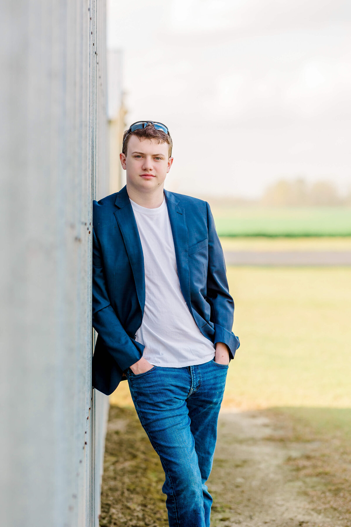 A person in a blue blazer, white t-shirt, and jeans leans against a wall outdoors. They have sunglasses on their head and their hands in their pockets. The background shows grass and a blurred landscape.