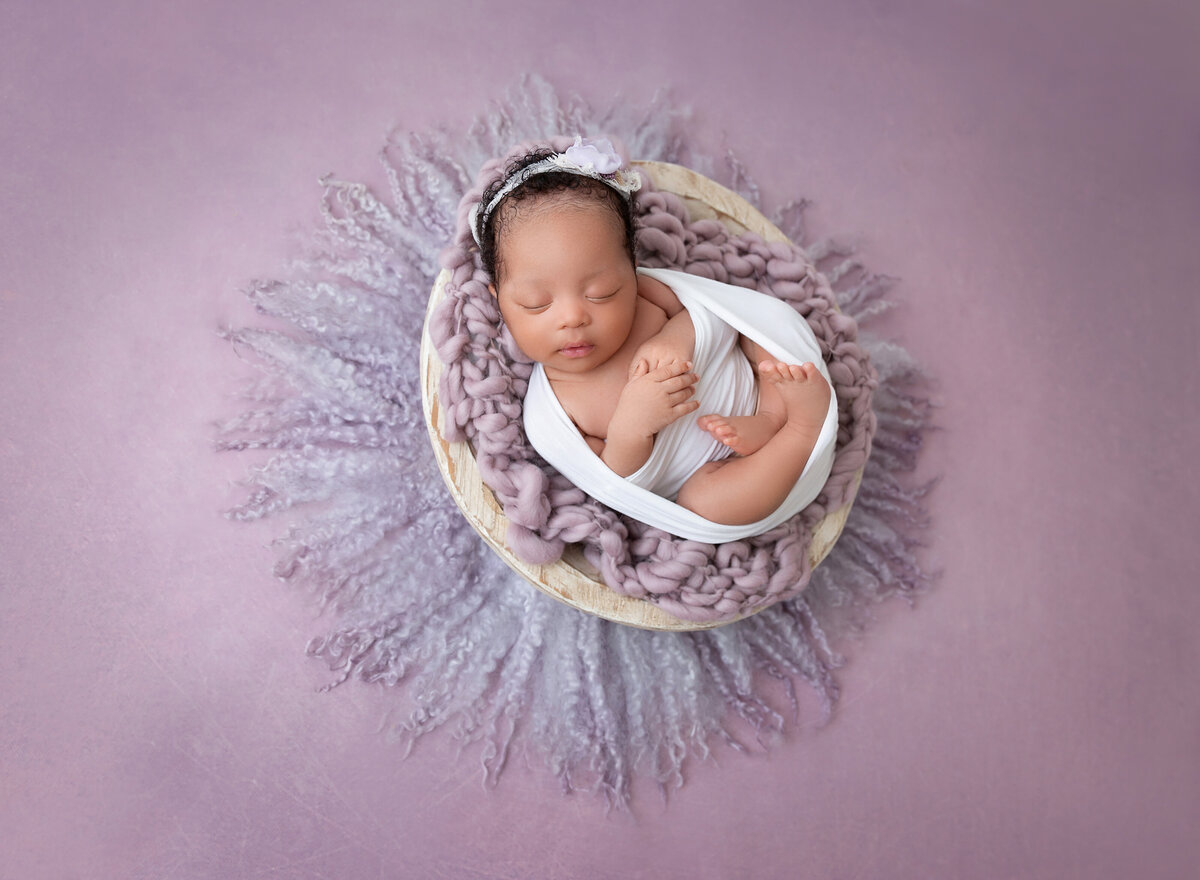 Newborn baby girl is wrapped in a white knit wrap with her hands and toes peeking out. She is sleeping in a basket covered with a purple knit blankets.