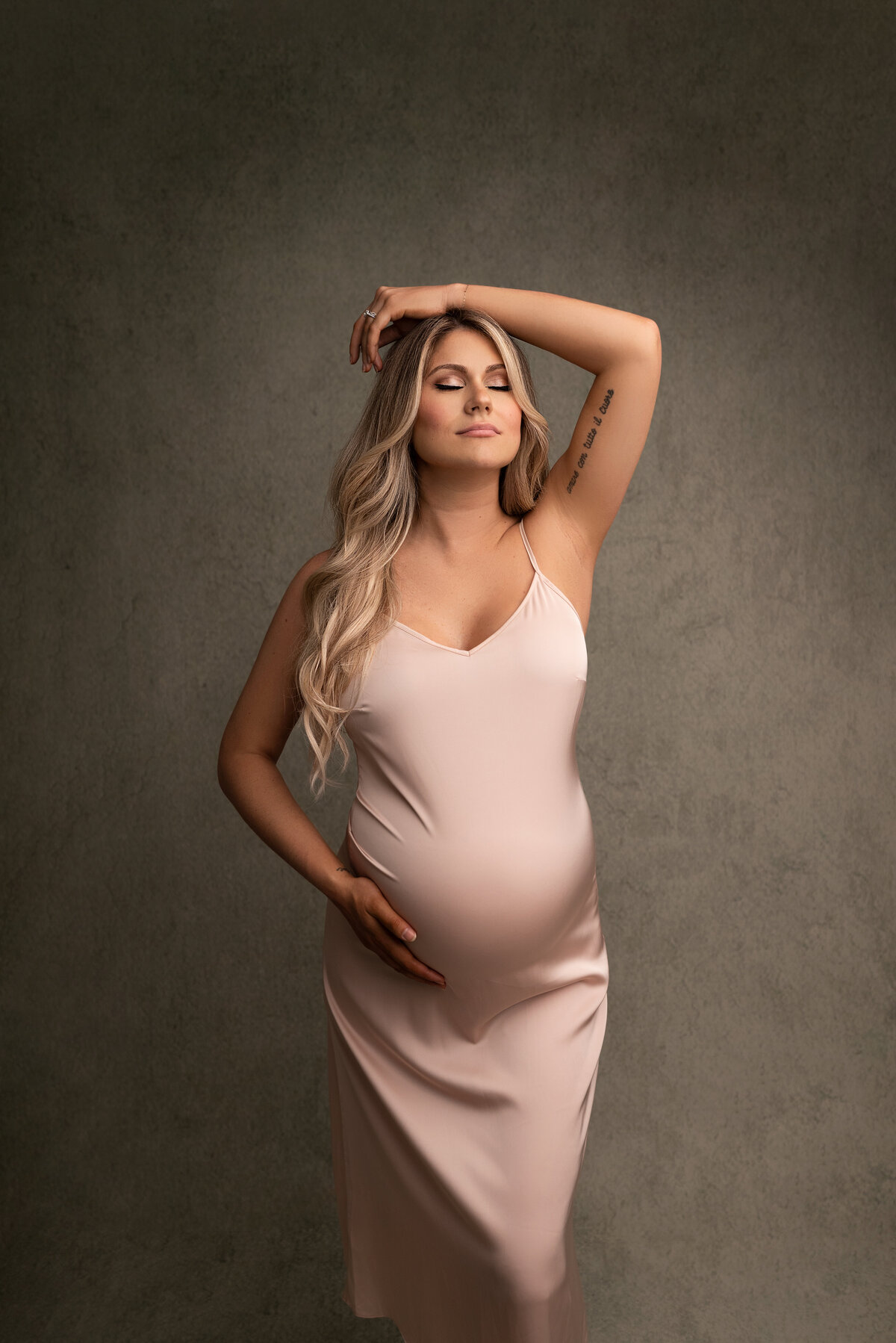 Philadelphia Main Line's best maternity photographer Katie Marshall captures expectant mom for a fine art maternity photoshoot. Woman in a long blush silky maternity slip is standing facing the camera. One hand rests to the side of her baby bump, the other arm is draped atop of her head. Her eyes are closed and chin tilted slightly up toward the light.