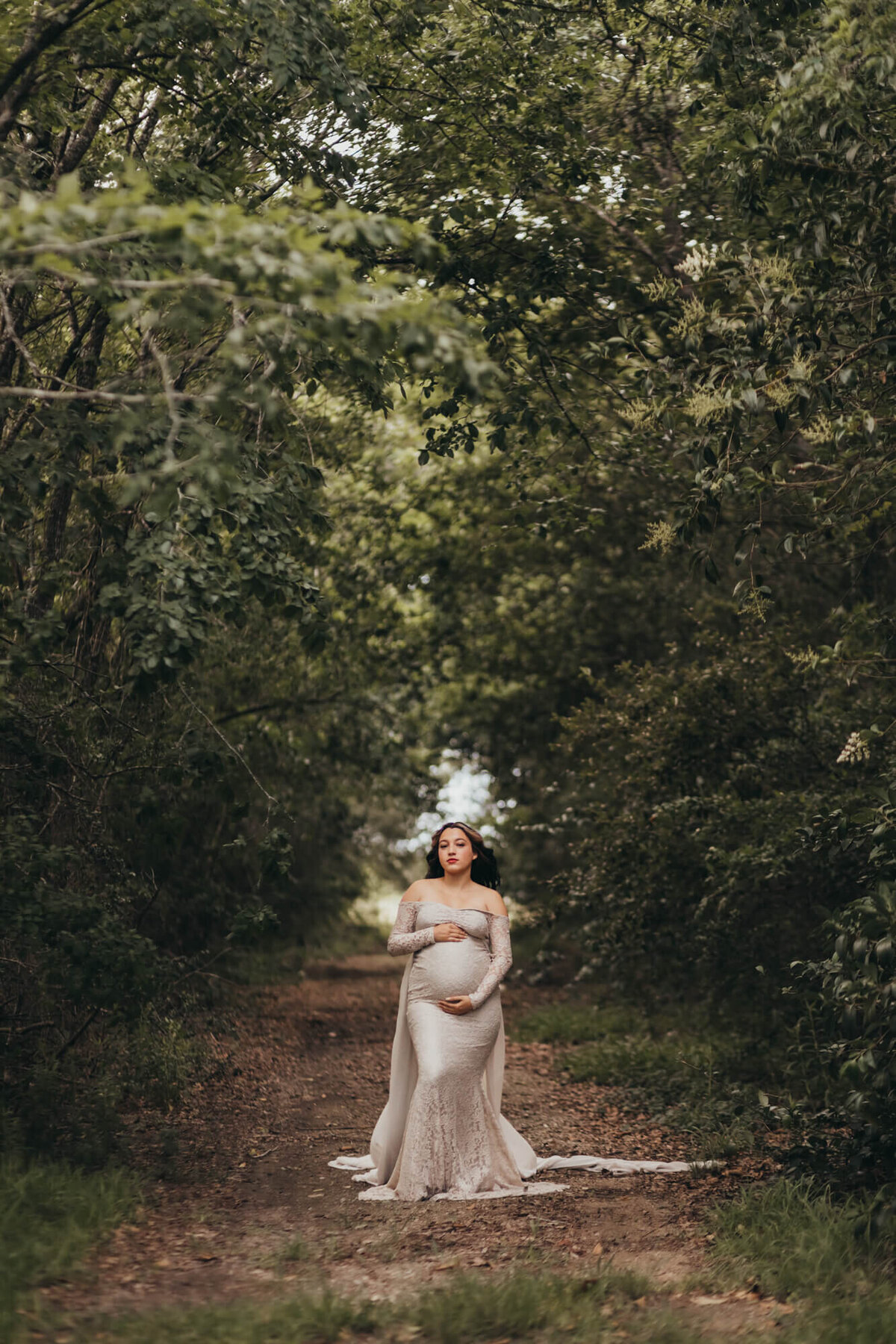 mom holds her belly in a tunnel of trees wearing a beige maternity gown