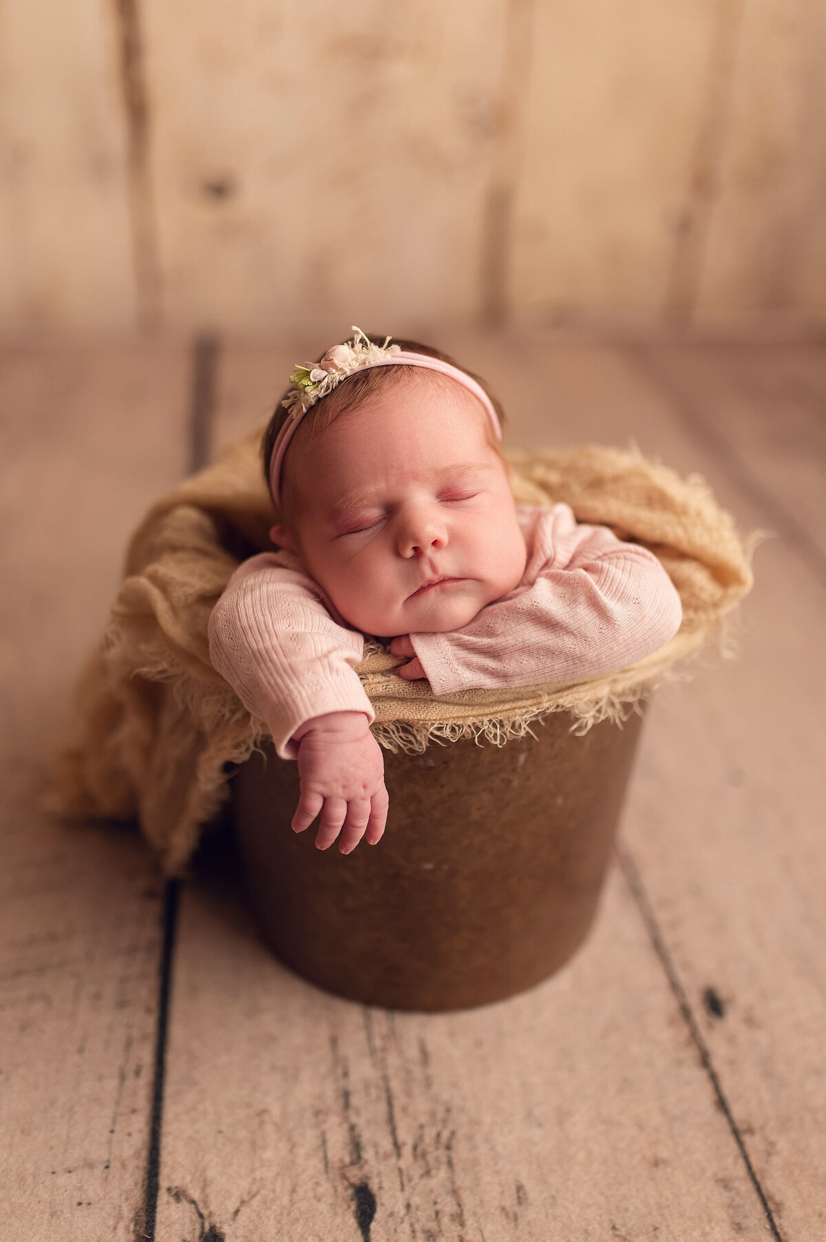 Portrait of baby girl asleep in a brown bucket filled with burlap-like fabric.  Infant was photographed in our Waukesha, WI studio.