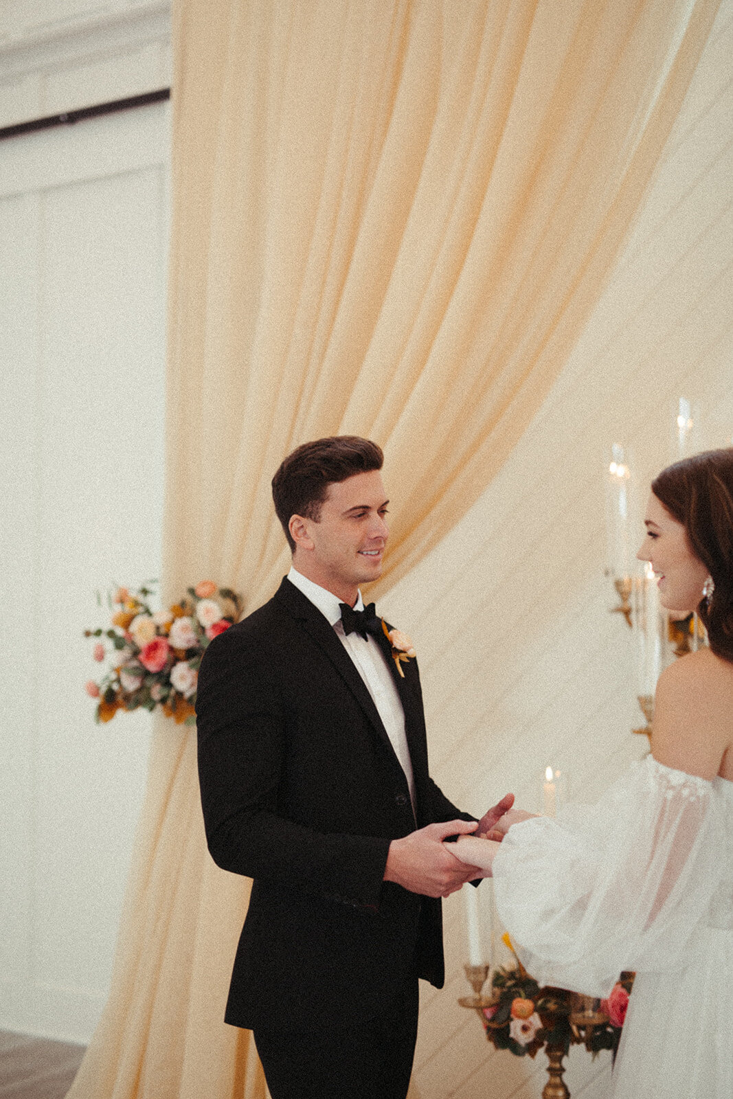 A groom wearing a black tuxedo holds bride wearing a white wedding gown by the hands in front of peach curtains.