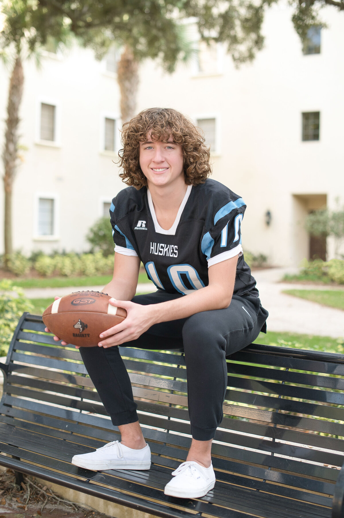 Hagerty High School senior boy in football jersey holding a football sitting on top of a bench smiling.