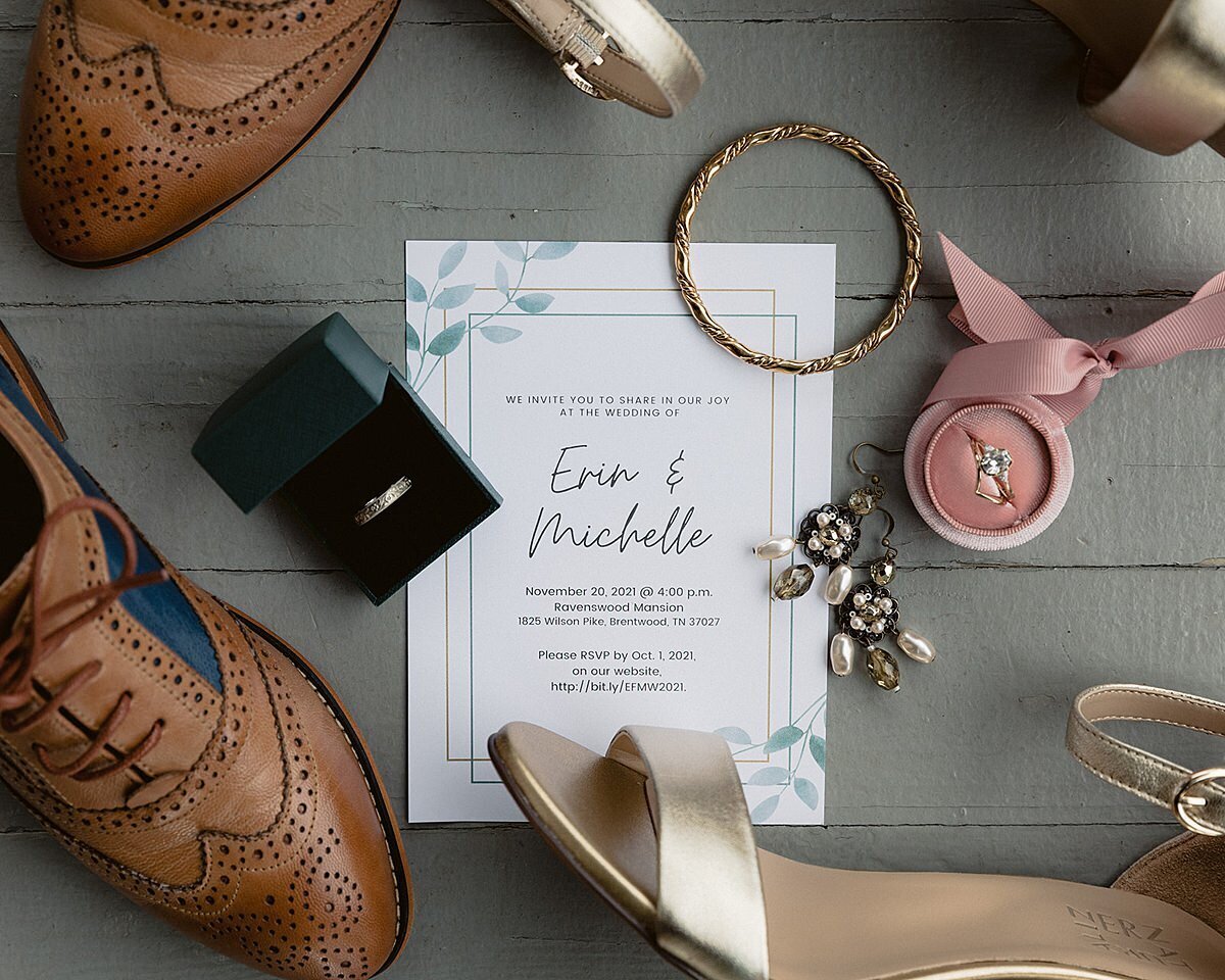 Brown wingtip shoes, gold strappy stilettos, pearl earrings and a gold bangle bracelet surround a wedding invitation with  watercolor leaves. A black ring box holds a plain platinum wedding band and another pink ring box holds a diamond engagement ring and diamond wedding band.