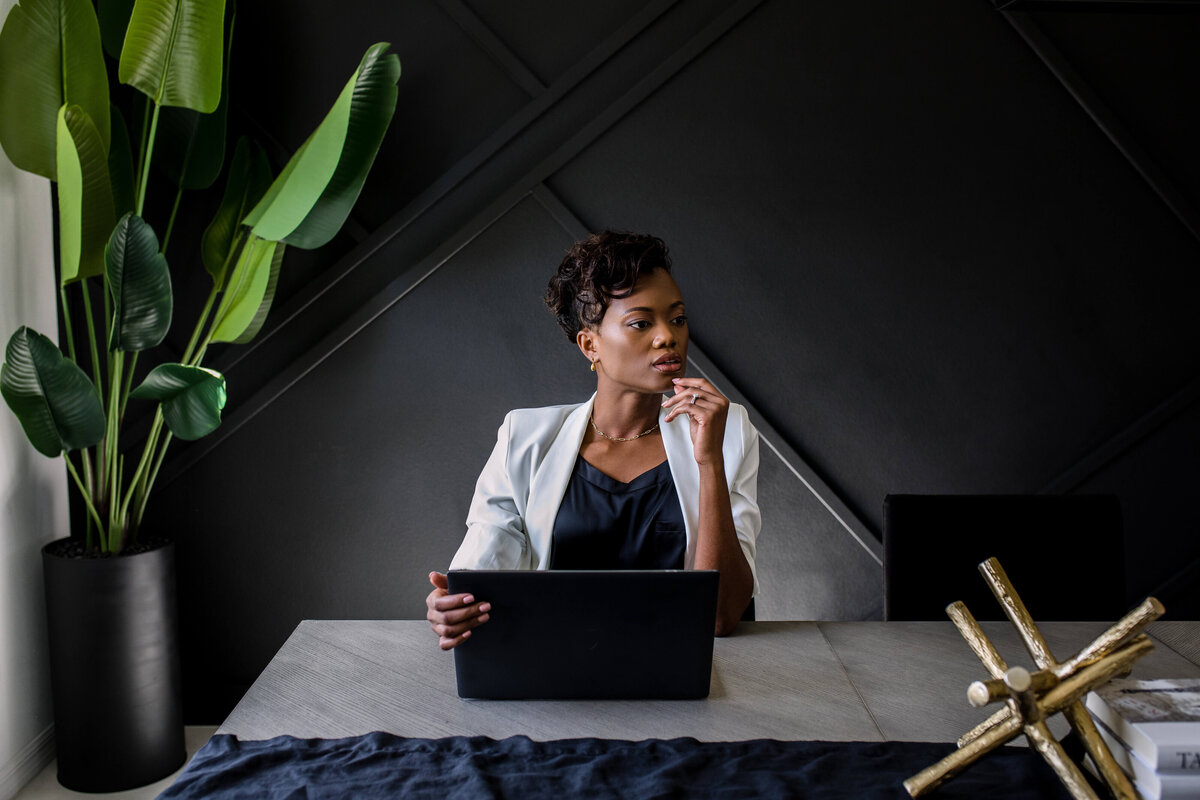 Brand photo of woman sitting at a desk with a black wall behind her holding her laptop and looking over her shoulder for a personal branding photo shoot with commercial photographers