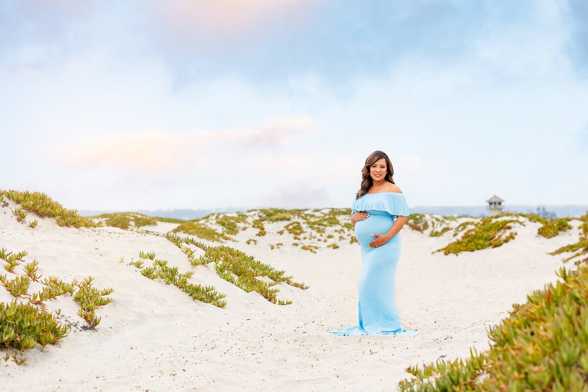 Woman in blue maternity dress holding her belly and posing in the sand dunes in Coronado