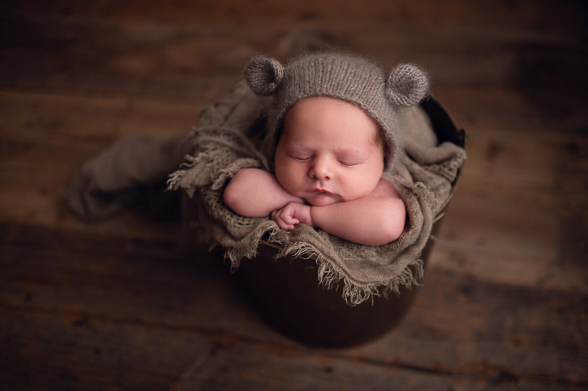 Newborn baby boy posed sleeping in a metal bucket  with woven neutral fabric around him in our Waukesha studio.  Infant is wearing a brown, knitted hat that has bear ears attached.
