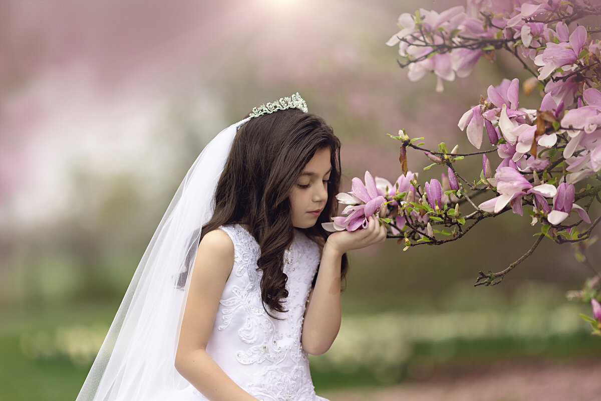 A young girl with dark hair smells some pink flowers while wearing a white communion dress and tiara