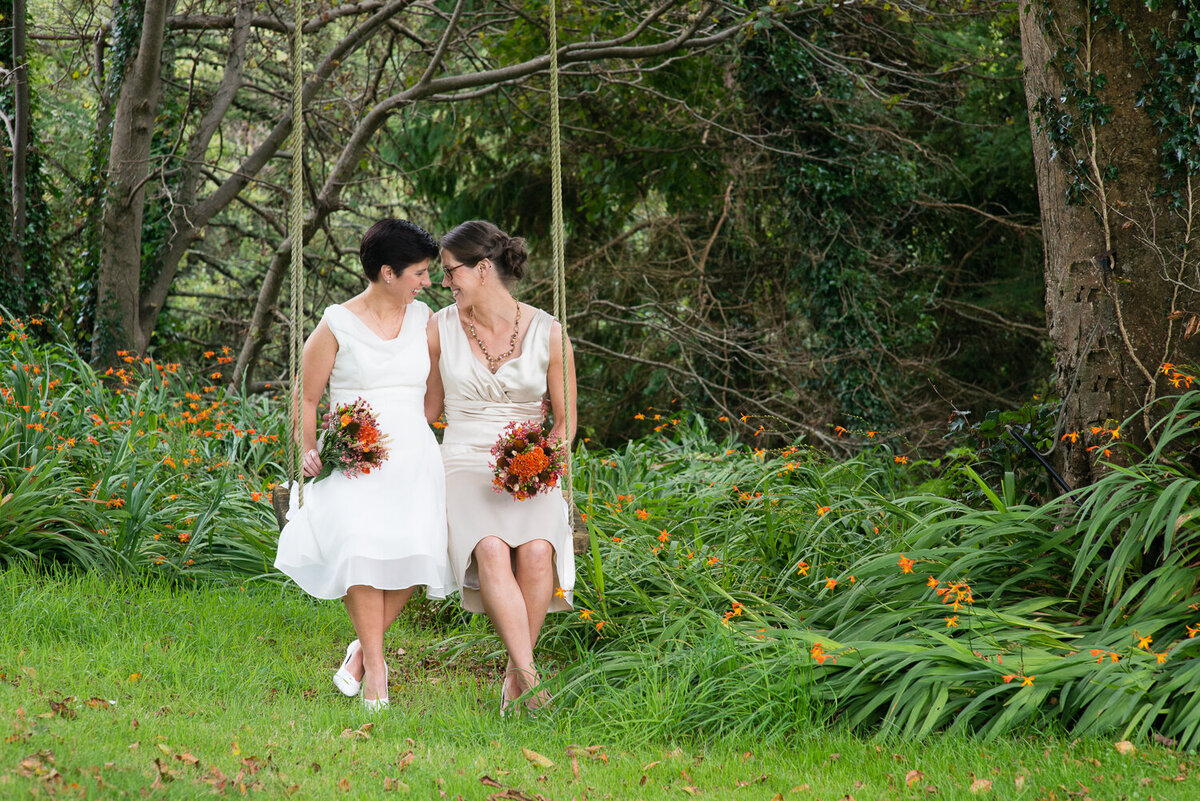 Gay wedding couple wearing white dresses, sitting on a swing, holding orange bouquets in a garden