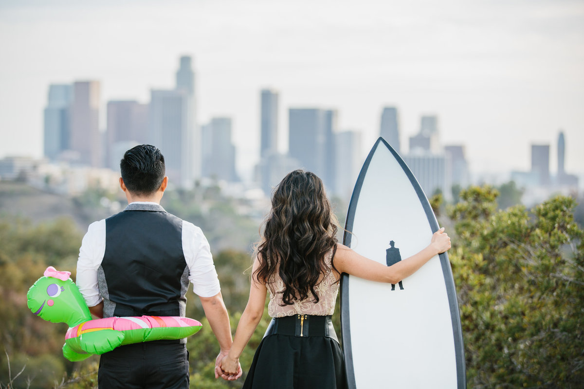 Couple holding hands looking at Los Angeles Skyline. She is holding a surfboard and he has a turtle pool float around his midsection