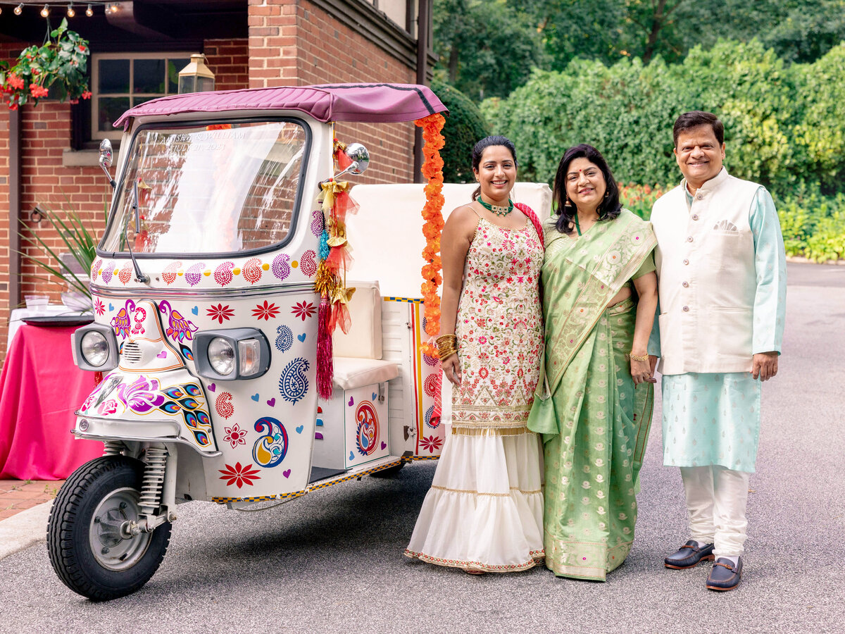 A smiling family of three stands beside a vibrantly decorated rickshaw. The woman in the middle wears a green sari, while the others are in traditional Indian attire. The setting is outdoors, with greenery in the background.