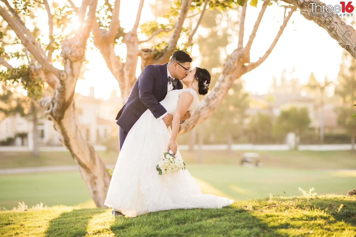 Groom dips and kisses his Bride under a tree during a photo shoot