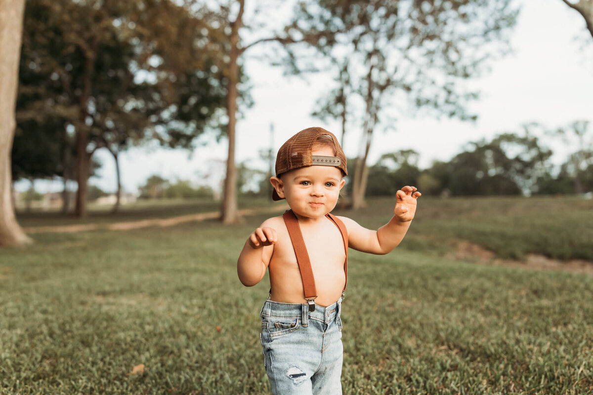 baby boy walks around park wearing a backwards cap and suspenders