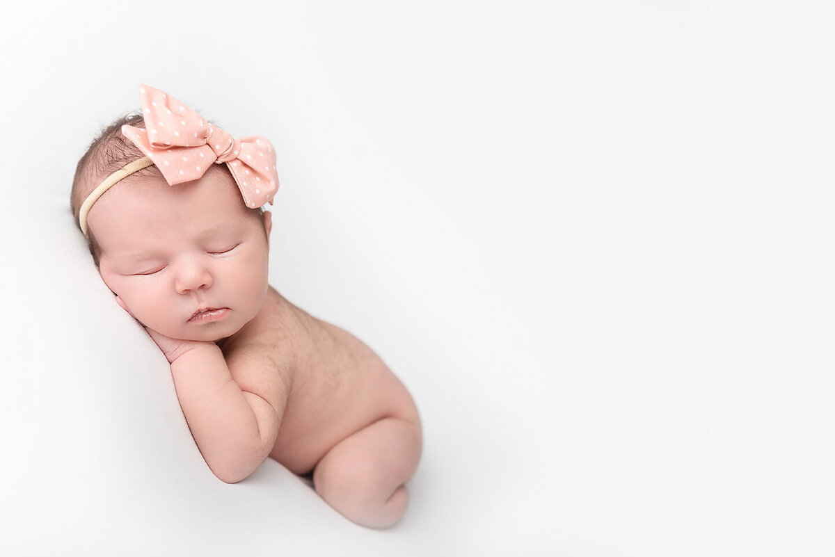 A sleeping newborn baby girl on a white pad in a pink bow