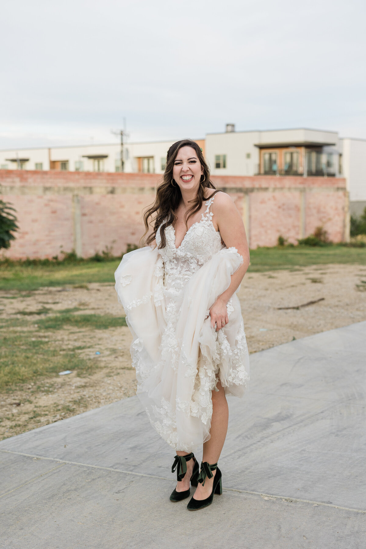 Portrait of a bride lifting up her wedding dress to show off her shoes while smiling fully after her wedding ceremony at The 4 Eleven in Fort Worth Texas. The dress is sleeveless, intricate, white, and long while the shoes are high heel, velvet, and green.