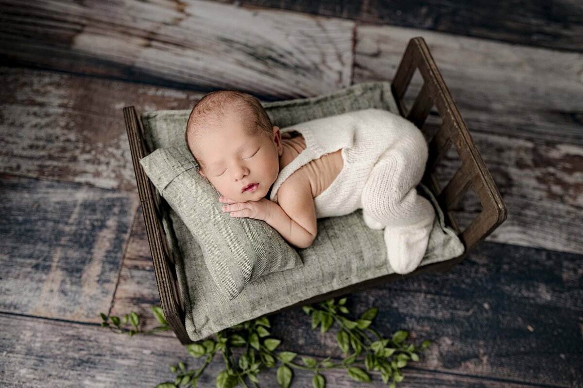 newborn baby boy dressed in cream knit overalls is posed on a bed with green  and brown accents
