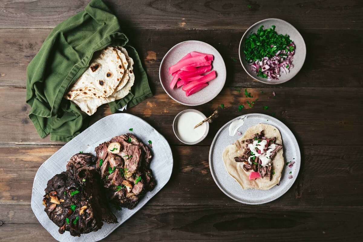 Omayah Atassi Dubai Food Photographer and Food Stylist - Overhead shot of shawarma-spiced lamb shoulder with homemade flatbread, pickled turnips, parsley, chopped red onion, and tahini sauce