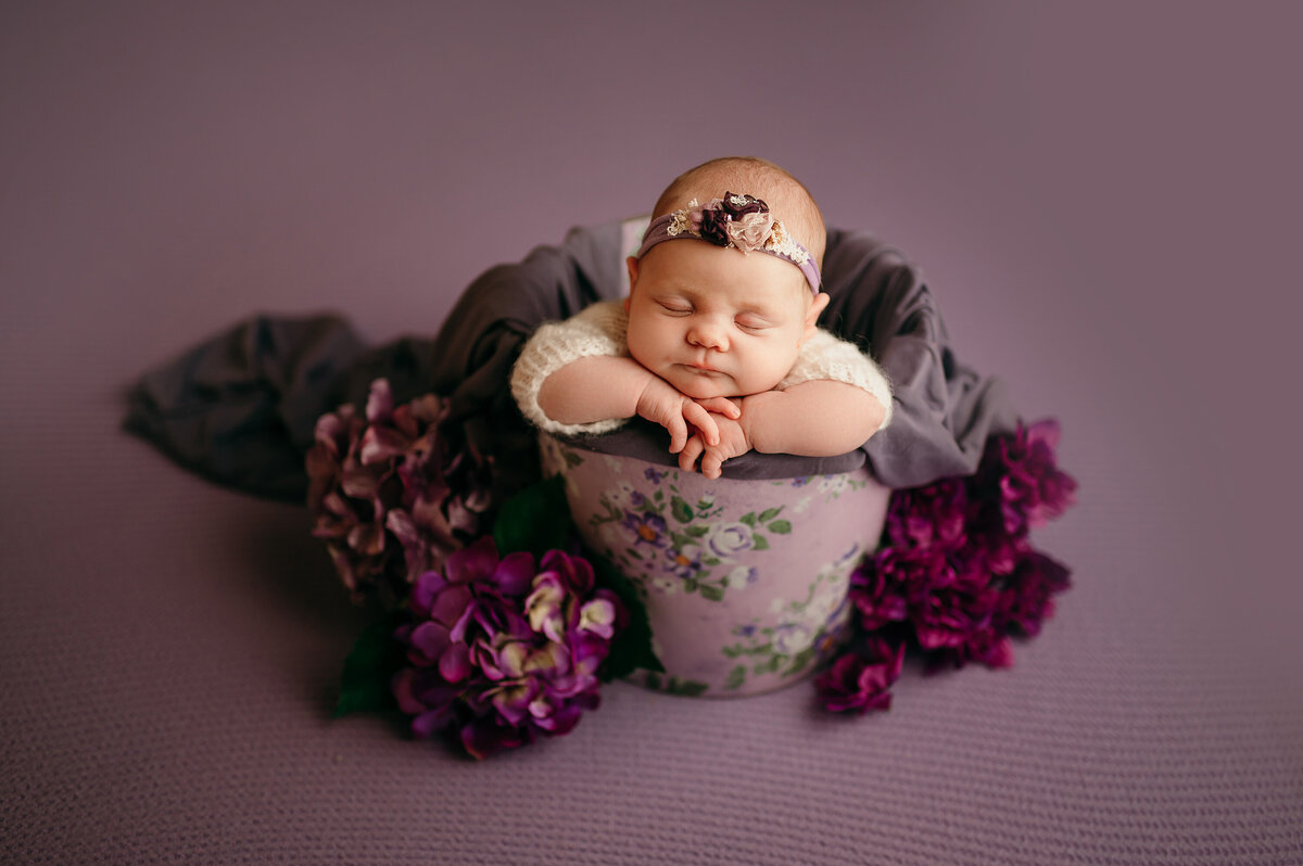 Portrait of a sleeping newborn in a bucket surrounded by purple florals. Baby is wearing a floral headband and ivory sweater.