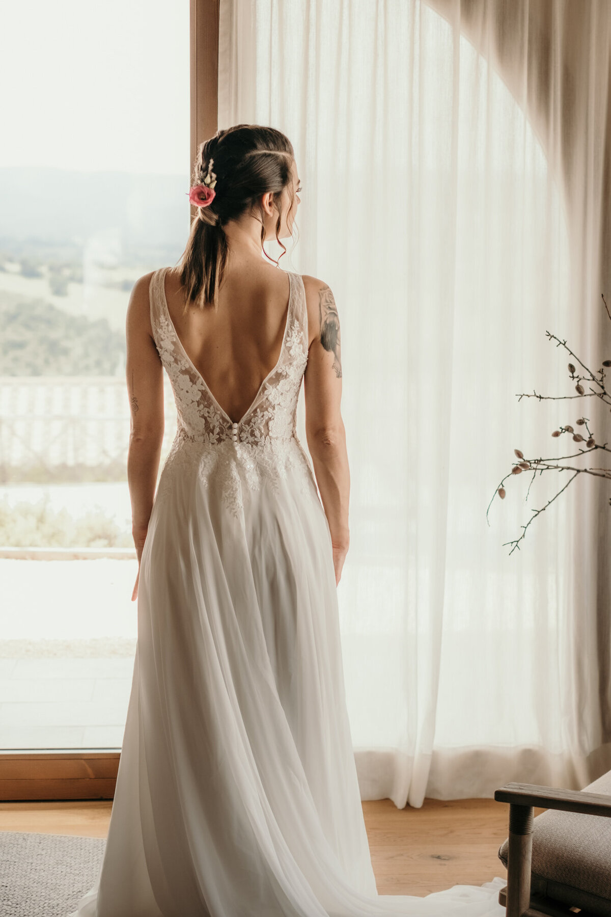 Fertig gestylt steht die Braut in Richtung Fenster blickend mit dem Rücken zur Kamera.