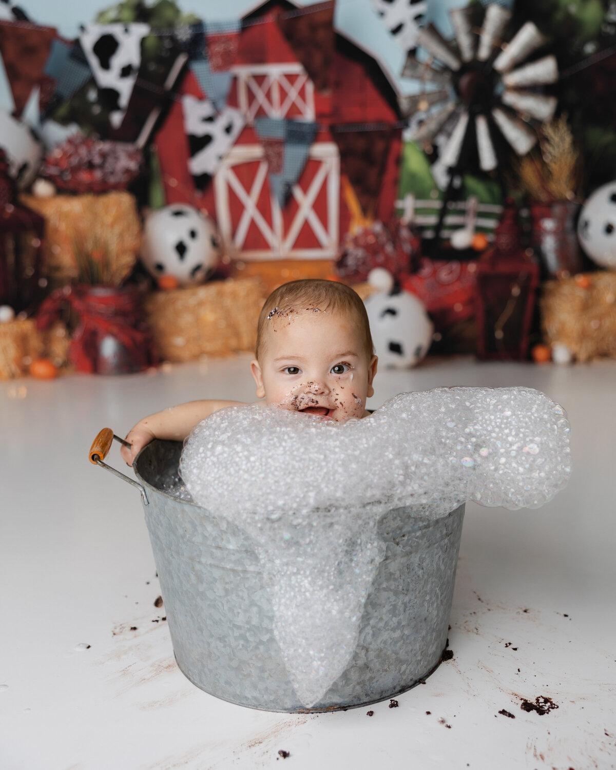 Smiling baby boy sitting in a bubble-filled tin tub after his farm-themed cake smash, with cake on his face and a barnyard backdrop behind him.
Photograph by Yucaipa cake smash photographer Kaitlyn Dawn Photography.