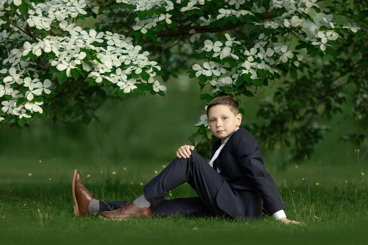 A young boy sits in a green lawn under a white flowering tree in a black suit for a New Jersey Communion Portrait Photographer