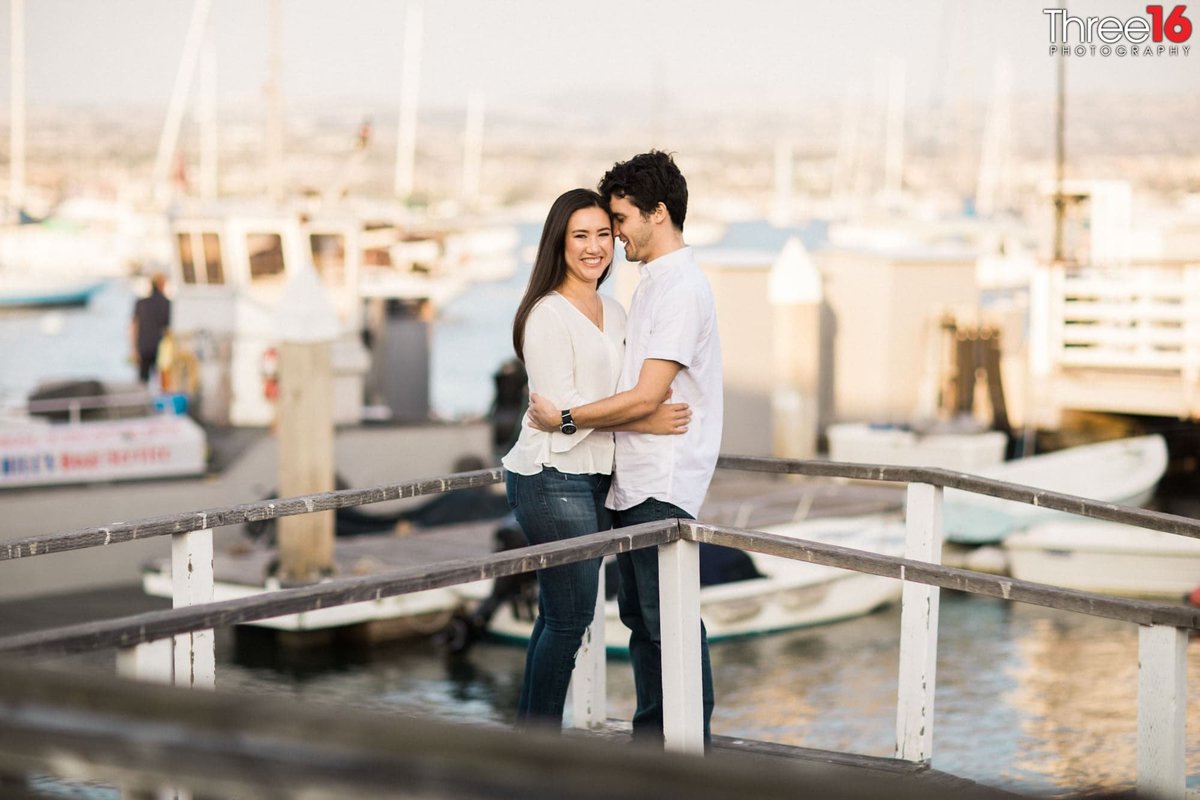 Engaged couple embrace each other as he looks at her while she smiles for the camera on the docks in Balboa