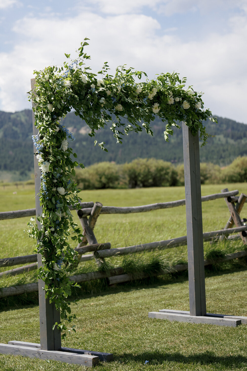 Wooden wedding arch decorated with lush blue and white florals, standing against the majestic backdrop of the Teton Mountains